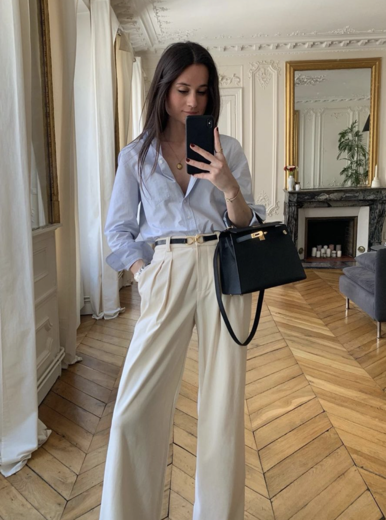 Woman wearing a light blue button-down shirt with wide-leg trousers, holding a black handbag, taking a mirror selfie in a stylish room.